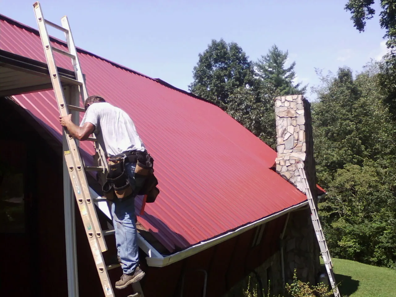 Roofer on a ladder beside a red metal roof and stone chimney — Soffit & Fascia Repair by Roofing Brothers in Archbald
